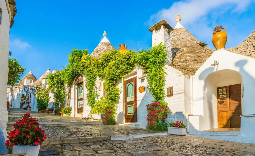 Ruas de pedra e casinhas tradicionais com telhados cônicos dos trulli em Alberobello, na região da Apúlia, Itália, cercadas por flores e plantas sob um céu azul vibrante.