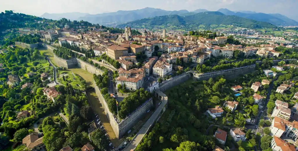 Vista aérea da Città Alta de Bérgamo cercada por muralhas venezianas, com construções históricas, vegetação abundante e montanhas ao fundo.