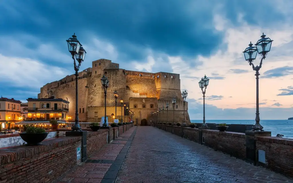 Vista do Castel dell’Ovo ao entardecer em Nápoles, Itália, com iluminação suave, céu dramático e o caminho de acesso ladeado por postes antigos à beira do mar.