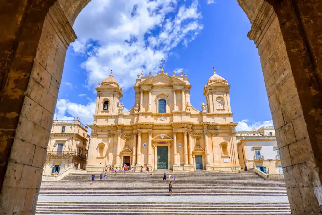 Fachada principal da Catedral de Noto, na Sicília, vista de frente por entre dois arcos de pedra. A escadaria leva até a entrada da igreja barroca, iluminada pelo sol sob um céu azul vibrante com nuvens.
