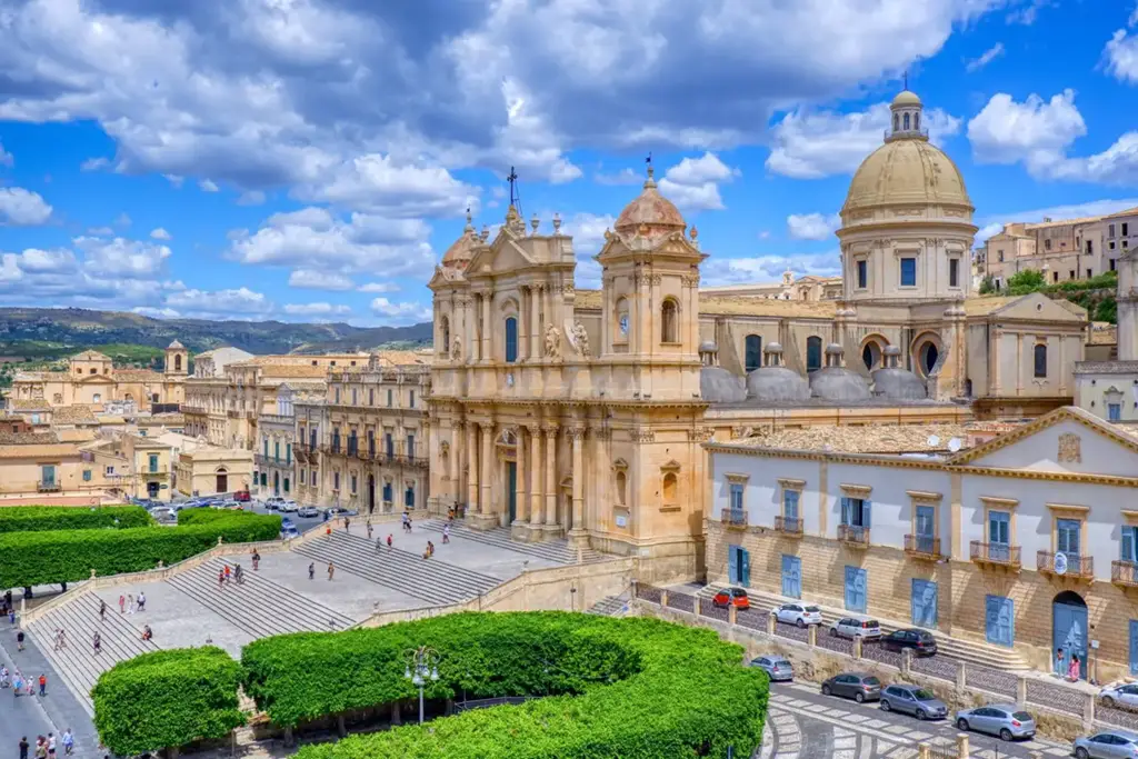 Vista da imponente Catedral de Noto, na Sicília, Itália, com sua fachada barroca em tom de areia sob um céu azul com nuvens. A escadaria em frente e a praça ao redor estão movimentadas com turistas.