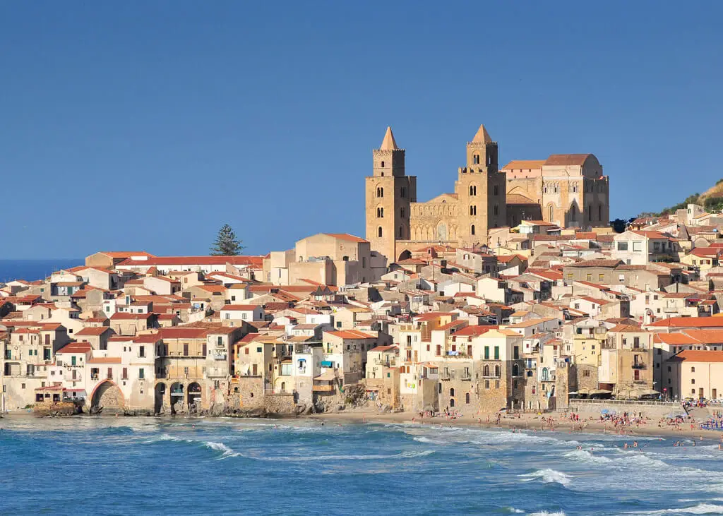 Vista panorâmica da cidade costeira de Cefalù, na Sicília, com suas casas de telhado vermelho agrupadas à beira-mar e a imponente Catedral de Cefalù ao fundo, em um dia ensolarado.