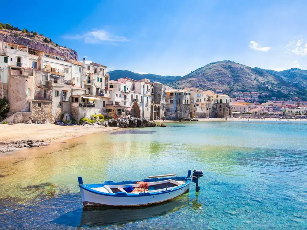Barco de pesca ancorado em águas cristalinas com vista para as casas à beira-mar de Cefalù, na Sicília, e colinas ao fundo sob céu azul claro.