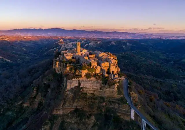 Vista aérea de Civita di Bagnoregio ao entardecer, vila medieval no topo de um penhasco cercado por vales, com acesso por uma longa passarela elevada.