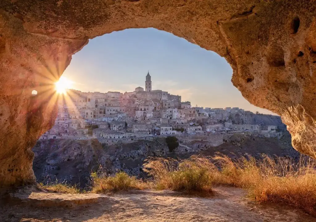 Vista de Matera ao pôr do sol, emoldurada pela entrada de uma caverna com vegetação seca em primeiro plano e o sol brilhando por entre as construções históricas de pedra ao fundo.