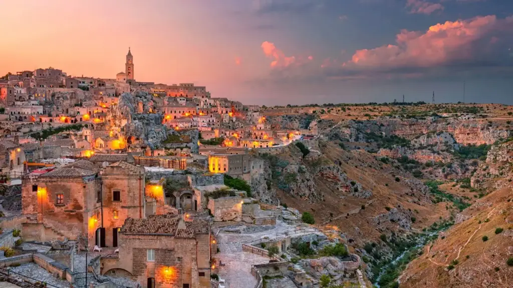 Vista panorâmica de Matera ao entardecer, com suas construções históricas esculpidas nas rochas iluminadas por luzes amareladas, contrastando com o céu rosado e o cânion ao lado.