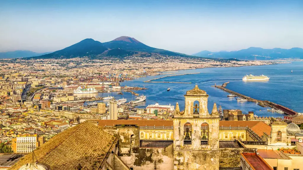Vista panorâmica de Nápoles, na Itália, com o imponente vulcão Vesúvio ao fundo, o porto movimentado com cruzeiros e navios, e construções históricas em destaque sob um céu claro.