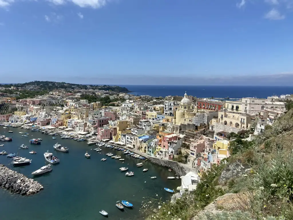 Vista panorâmica da ilha de Procida, na região da Campânia, Itália, com suas casas coloridas à beira-mar, barcos ancorados no porto e o mar azul profundo ao fundo sob um céu claro.
