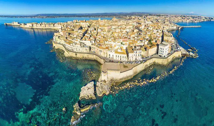 Vista aérea da ilha de Ortigia, centro histórico de Siracusa, na Sicília, cercada por águas azul-turquesa e com construções históricas à beira-mar.