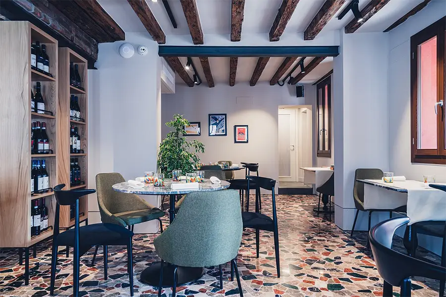 Sala aconchegante do restaurante Wisteria, em Veneza, com piso de pedras coloridas, teto com vigas de madeira e mesas elegantes
