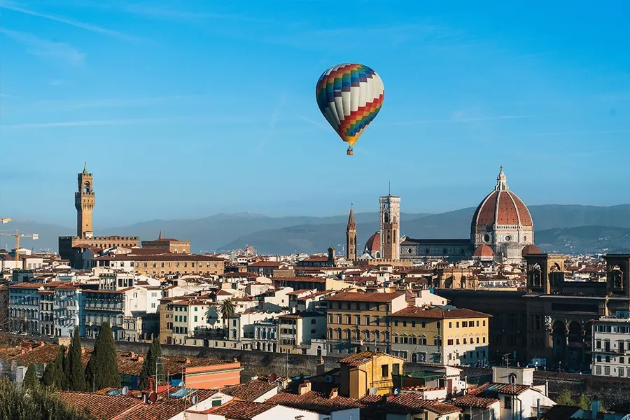 Balão de ar quente sobrevoando Florença, com vista panorâmica do Duomo, torres medievais e montanhas ao fundo em um céu azul claro.
