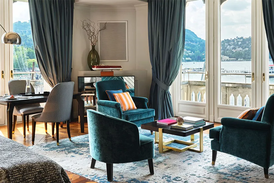Sala de hotel luxuosa com vista para o Lago di Como, com decoração elegante em tons de azul, poltronas aconchegantes e luz natural. 
