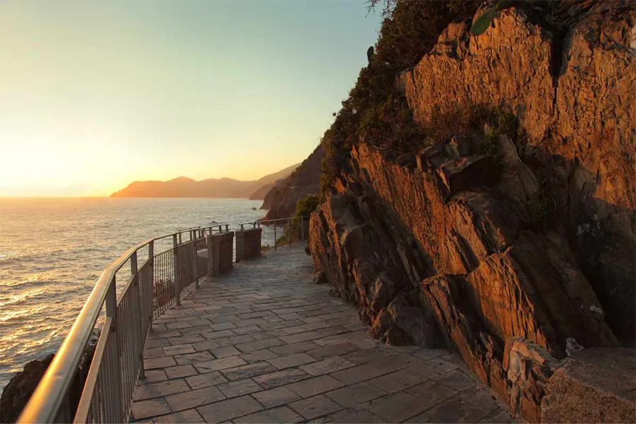 Trilha da Via dell’Amore em Cinque Terre, com vista para o mar ao entardecer e paredão rochoso iluminado pela luz dourada do pôr do sol. 