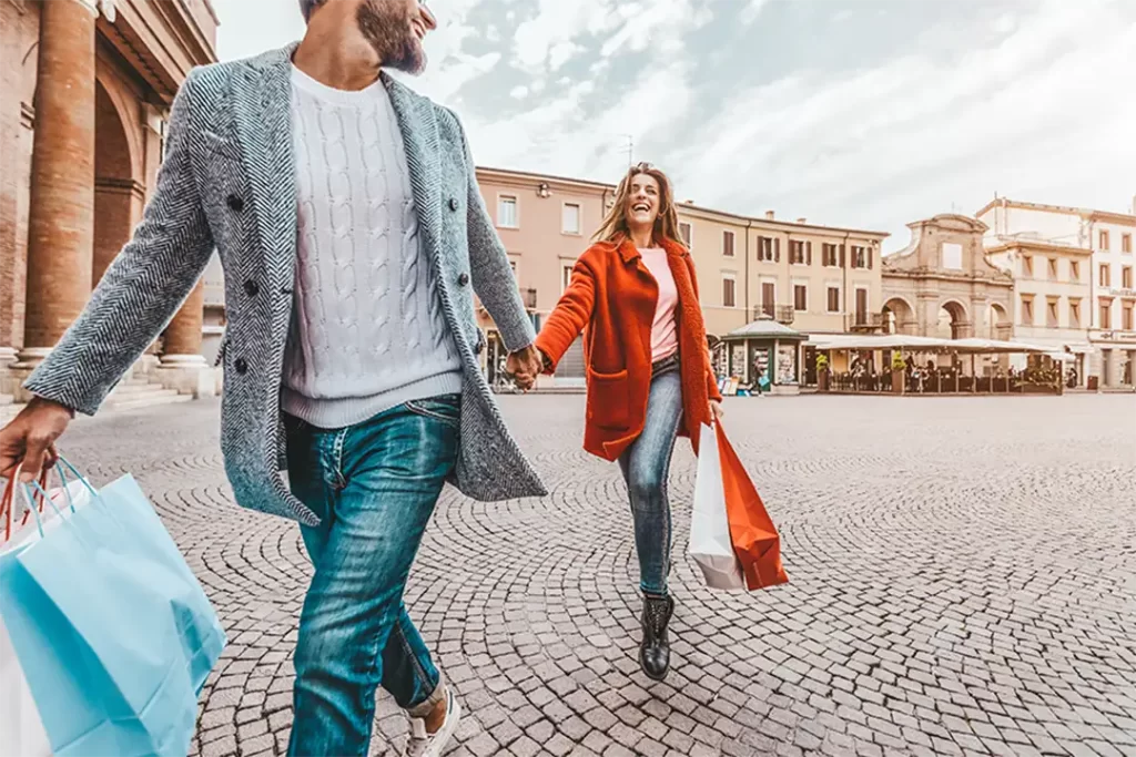 Casal sorridente com sacolas de compras em uma praça italiana, celebrando o estilo descontraído e atual da Moda Italiana.