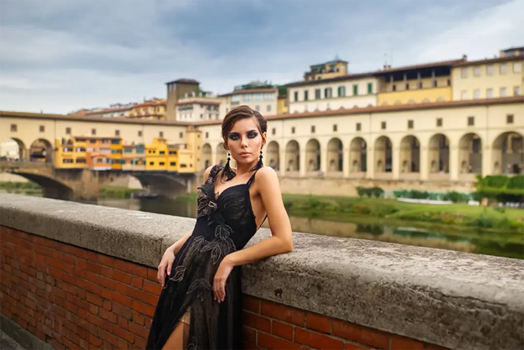 Modelo com vestido preto elegante posando em frente à Ponte Vecchio, unindo arte, história e sofisticação da Moda Italiana.