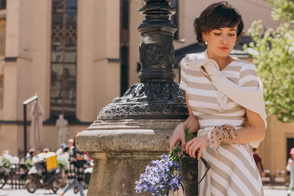 Mulher com vestido listrado e flores em mãos em rua clássica italiana, representando charme e feminilidade da Moda Italiana.
