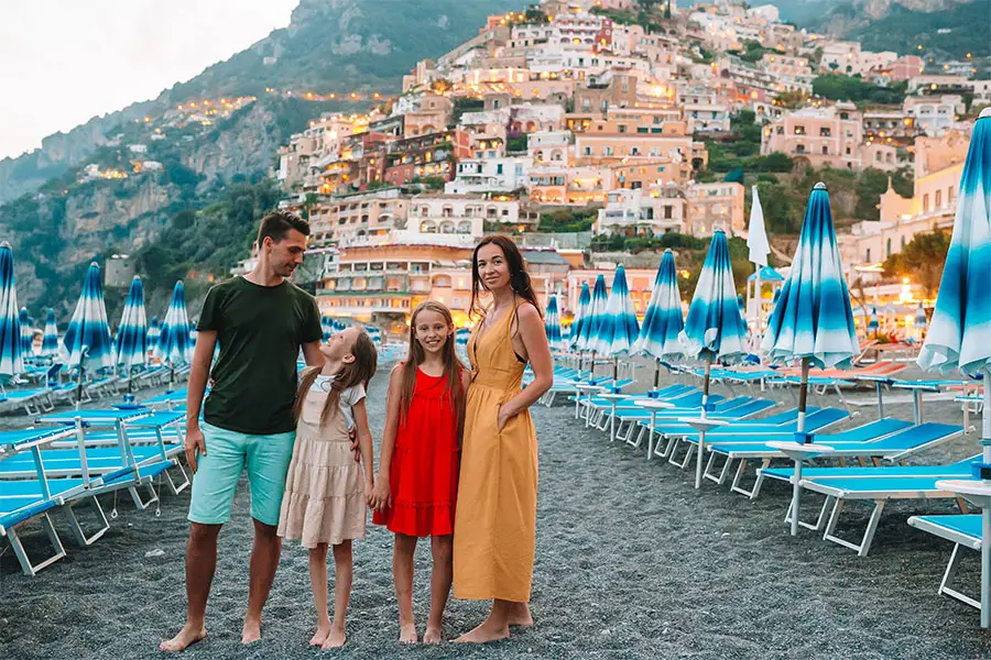 Família posando na praia de Positano, na Costa Amalfitana, cercada por guarda-sóis azuis e as charmosas casas coloridas na encosta ao entardecer, durante uma viagem em família para a Itália.