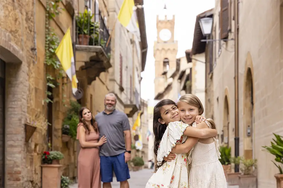 Duas meninas se abraçam sorrindo em uma rua charmosa de uma cidade medieval da Toscana, com os pais ao fundo durante uma viagem em família para a Itália.