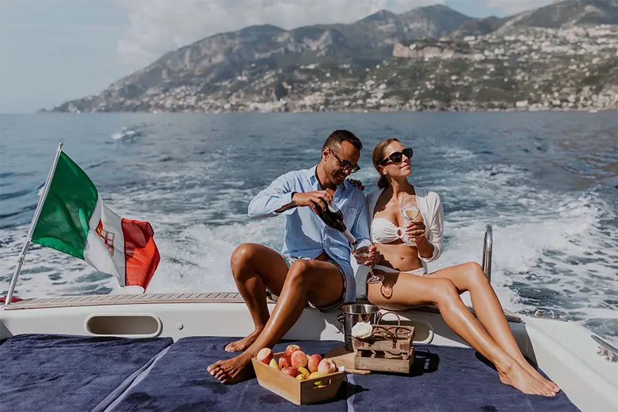 Casal aproveitando passeio de barco na Itália com taças de vinho a bordo de um iate, navegando pela Costa Amalfitana com paisagem montanhosa ao fundo e bandeira italiana ao vento.