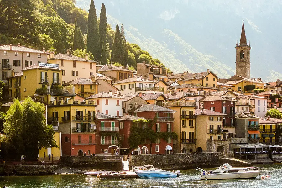 Vilarejo à beira do Lago de Como com barcos atracados no cais, cenário ideal para passeios de barco na Itália entre montanhas e arquitetura charmosa.
