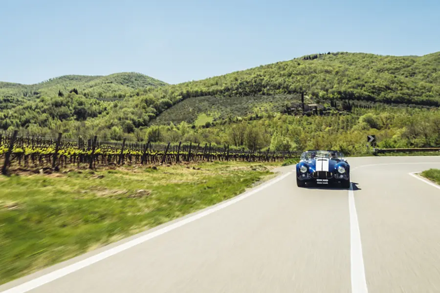 Carro esportivo clássico dirigindo por estrada sinuosa entre vinhedos e colinas verdes na rota Chiantigiana, Toscana, Itália