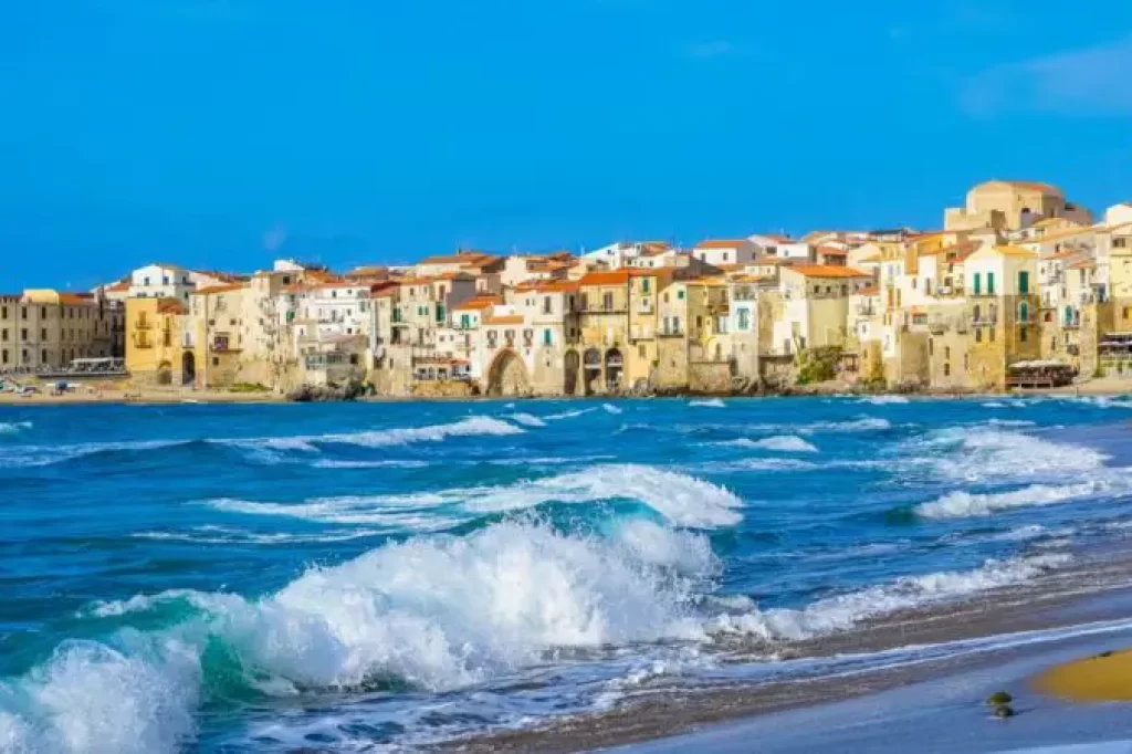 Vista panorâmica da cidade costeira de Cefalù, na Sicília, com casas históricas à beira-mar e ondas quebrando na praia sob um céu azul.
